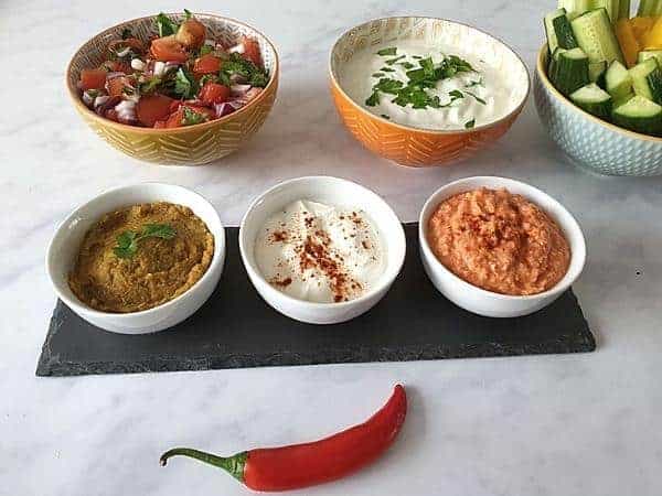 selection of dips in white bowls on a slate platter on marble tabletop with larger colourful bowls of more dips