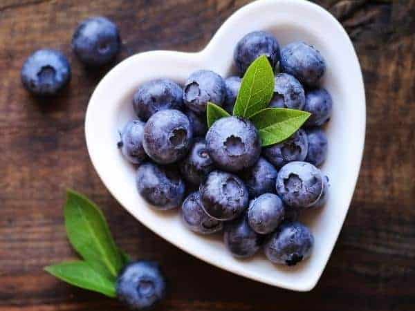 A heart-shaped bowl filled with blueberries and two green leaves sits on a wooden surface, reminiscent of the natural ingredients often found in refreshing low-calorie cocktails.
