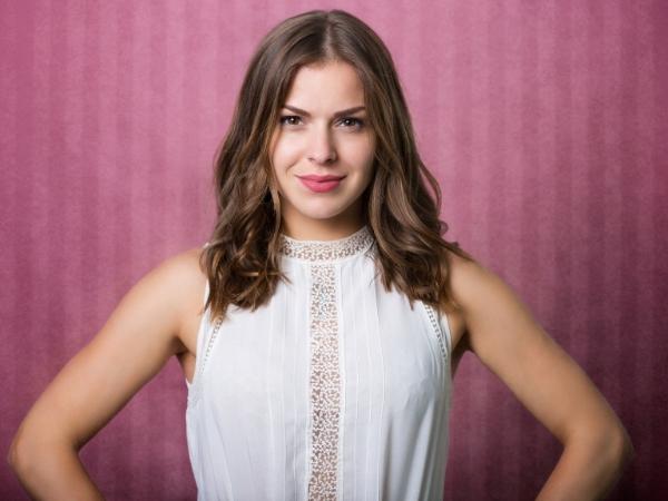 Brunette facing camera standing in front of a pink background with her hands on her hips