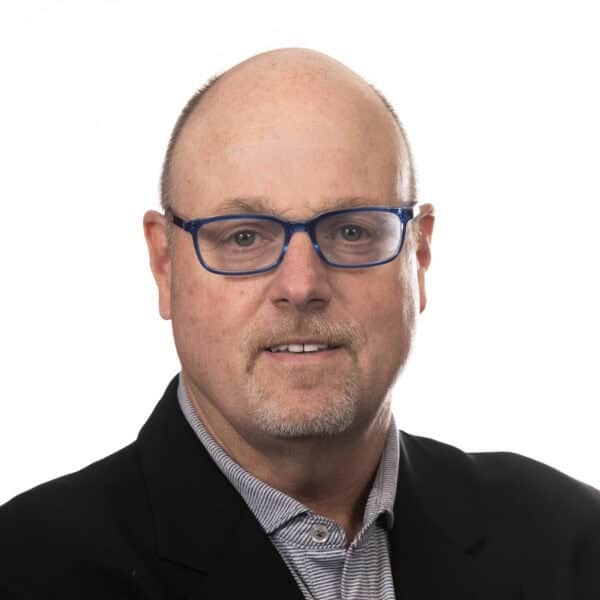 A middle-aged man with a bald head, wearing blue-rimmed glasses, a striped collared shirt, and a black jacket, poses in front of a plain white background.
