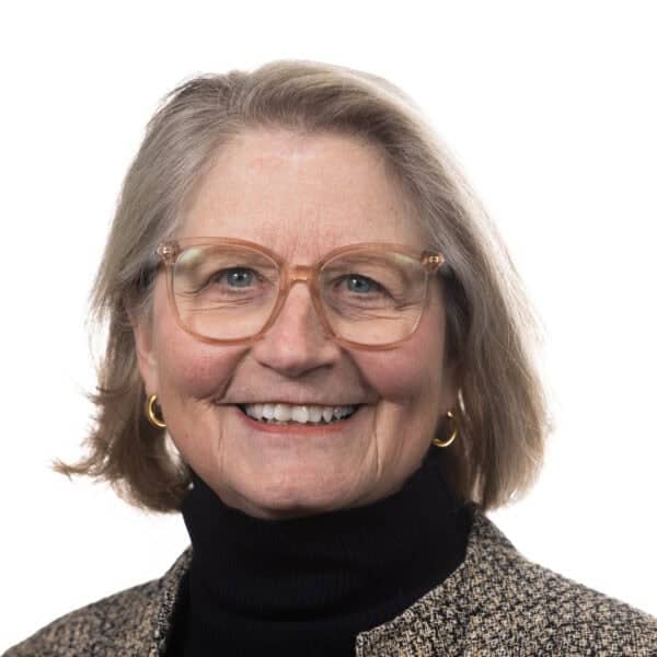 A smiling older woman with gray hair, wearing large clear glasses, gold hoop earrings, a black turtleneck, and a textured jacket, poses in front of a plain white background.