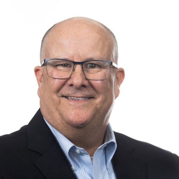 A middle-aged man with glasses, short hair, and a friendly smile is wearing a dark blazer over a light blue checkered shirt. He is pictured against a plain white background.