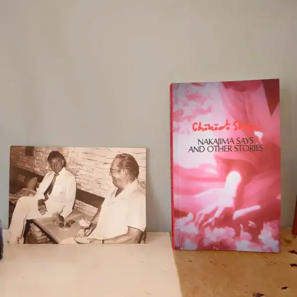 Bright pink and white book cover titled "Nakajima Says and Other Stories" by Chaiyo S. on a wooden surface with a vintage sepia photograph of two men sitting against a brick wall.