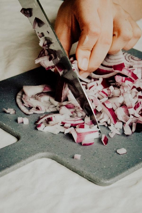 A person chops a red onion on a gray cutting board with a knife, preparing ingredients for delicious courgette bakes.