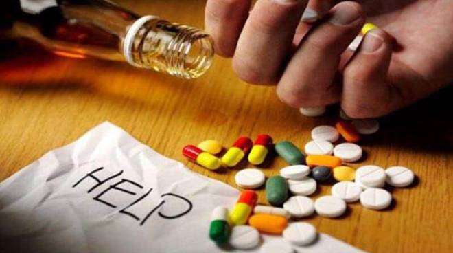 Caucasian hand, palm upwards nexxt to a tipped over bottle of whiskey which is full and open, and a 20 mixed, nondescript tablets on top of a white piece of paper with the word "HELP"written on it in capital letters in pencil.