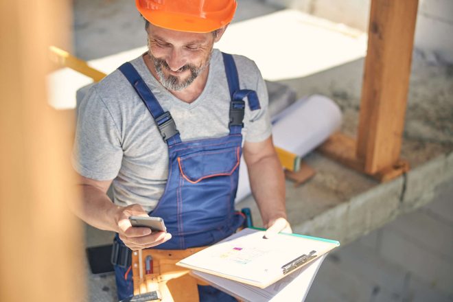 Smiling man with blueprints in his hands using his mobile phone in a half-finished building