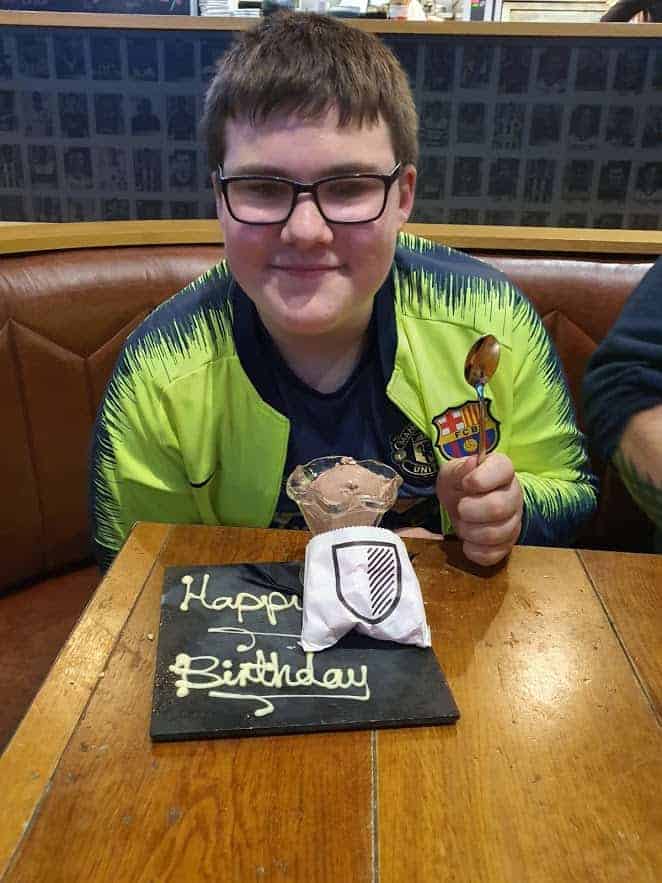 A young boy sitting at a table, smiling, with a happy birthday cake in front of him