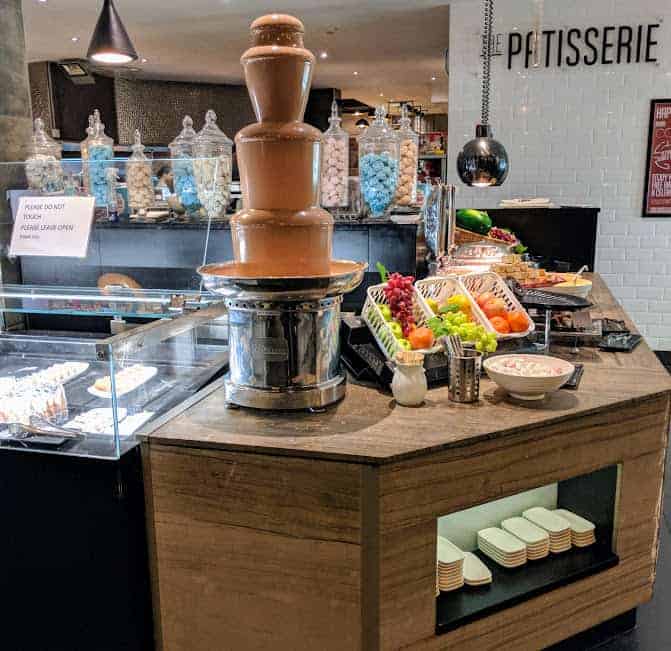 a chocolate fountain flowing with milk chocolate with fruit and cakes in background