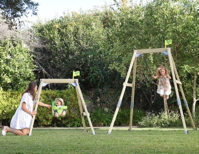 Two children playing on wooden swings in a green garden with grass and bushes.