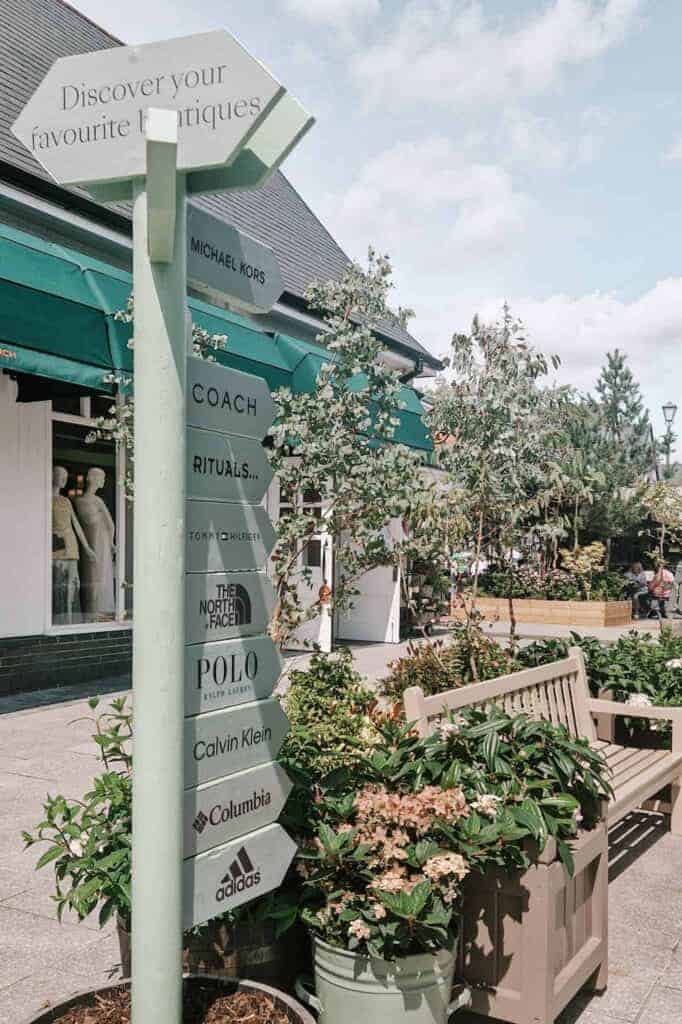 A directional sign at Kildare Village listing various high-end boutique names such as Michael Kors, Coach, and Polo Ralph Lauren, set against a background of shops with green awnings and potted plants.