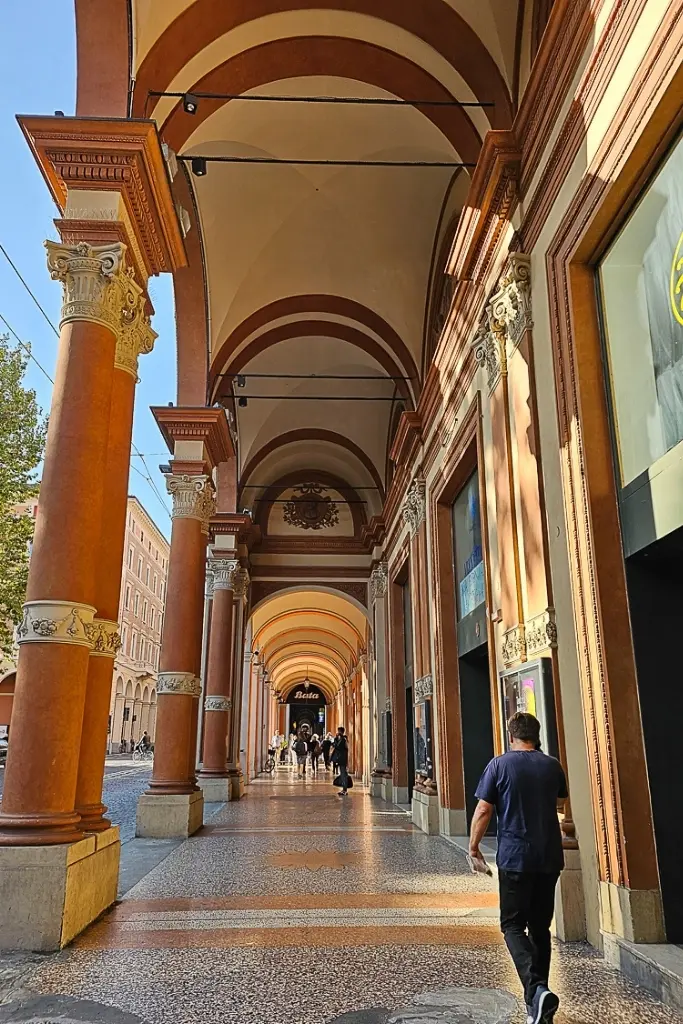 People walking under the elegant portico of Via Galliera in Bologna, Italy. The arched colonnade features detailed capitals and ochre-red columns, a quintessential example of Bologna’s UNESCO-listed portico architecture that stretches for kilometers throughout the historic center.
