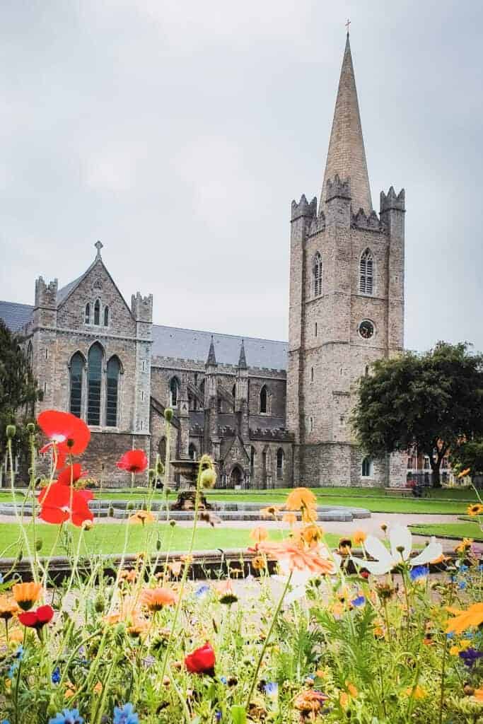 A historic stone cathedral with a towering spire, surrounded by a park with vibrant red and orange flowers in the foreground.