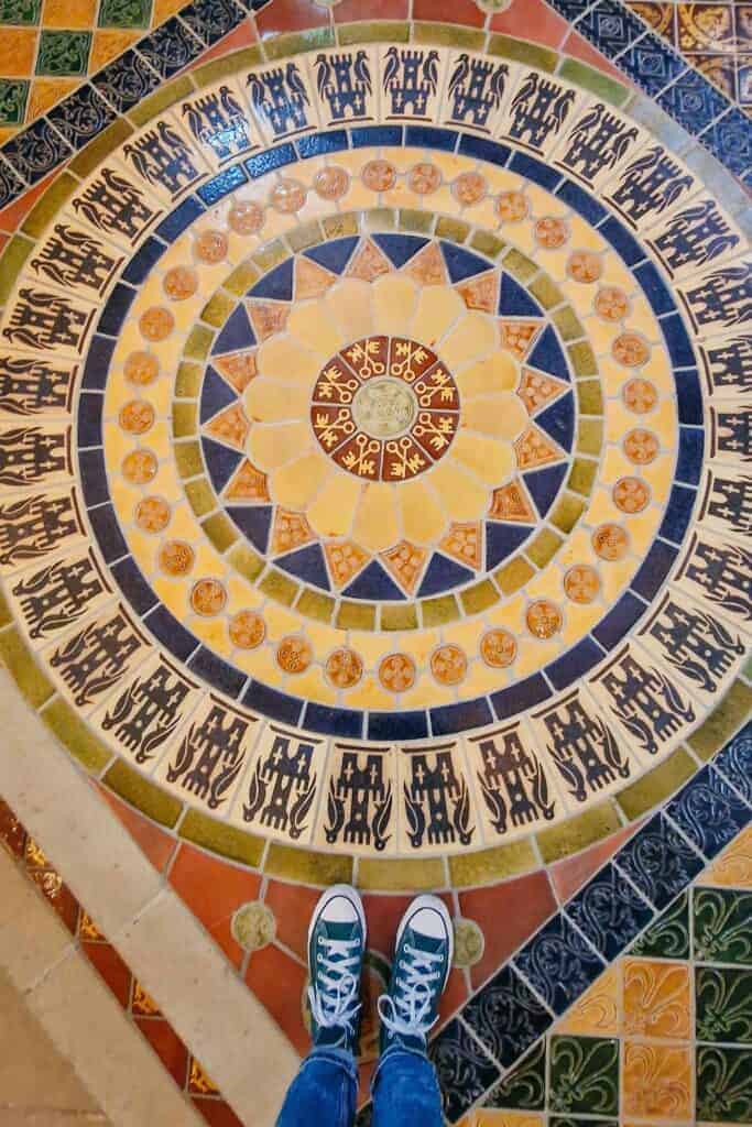 A detailed shot of the colorful mosaic floor inside the chapel at Swords Castle, featuring intricate patterns of interlocking designs in shades of yellow, blue, green, and red. The photographer's green sneakers are visible at the bottom of the frame.