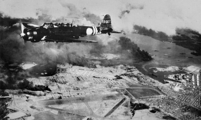 Japanse B5N2-bommenwerper boven Hickam Field tijdens tweede aanvalsgolf; USS Arizona brandt op de achtergrond.
