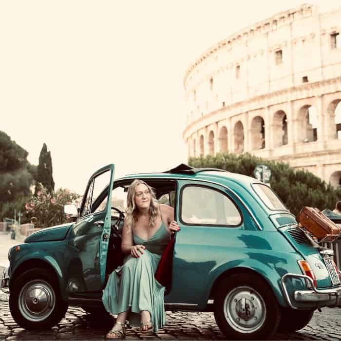 A woman in a light green dress sits in the doorway of a dark green vintage fiat 500 car in front of Rome's Colosseum at dawn, illuminated by the early morning golden light. She's Natalie, from Travel More With Nat.