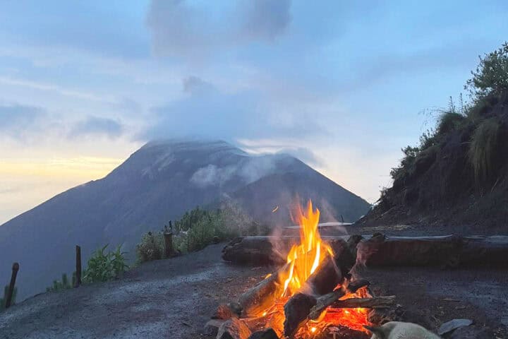 Campfire glowing at night on Acatenango campsite with a dog resting beside the fire and Fuego volcano in background