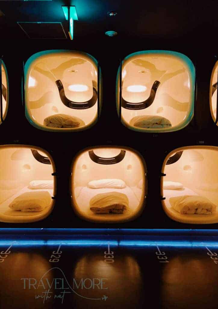 A sleek row of glowing capsule pods stacked two high in a dimly lit room, each with a neatly made bed and ambient lighting. Capsule hotels offer a futuristic, compact option for solo female travellers who value privacy, security, and affordability in urban stays.