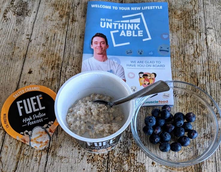 fuel salted caramel porridge pot with bowl of blueberries and on a wooden table with booklet and spoon in the porridge