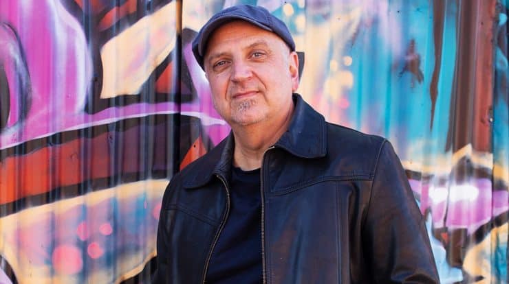 Older man in black leather jacket standing in front of colorful graffiti wall, urban street style, confident expression, music industry professional, artist, creative, street art atmosphere, hypebot content.