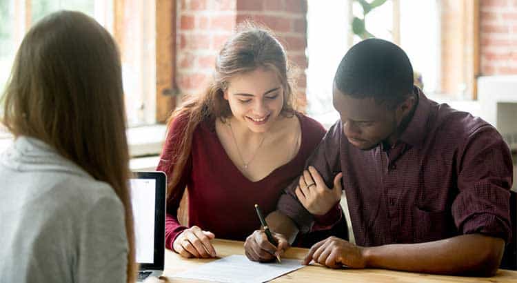 Lifestyle image of a smiling couple signing real estate documents during a meeting in a bright, modern office with exposed brick accents.