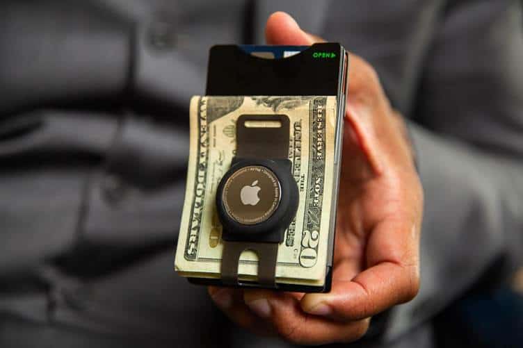 A person is holding a slim wallet containing credit cards and a stack of U.S. twenty-dollar bills. Attached to the wallet is a keychain with a device featuring an Apple logo. The person is wearing a dark-colored long-sleeve shirt.