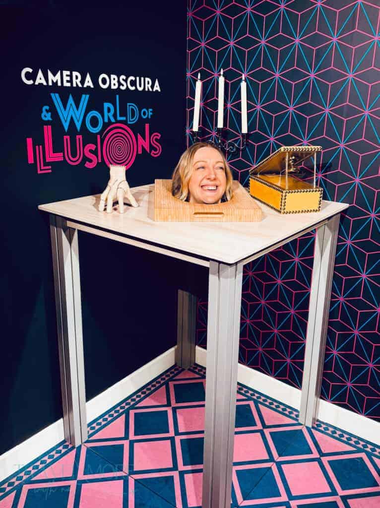 Visitor posing in a playful optical illusion exhibit at Camera Obscura in Edinburgh, with neon geometric walls and a “World of Illusions” sign. The perfect wet weather activity for a Edinburgh long weekend itinerary.