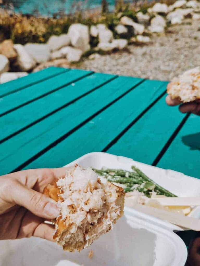 A close-up of a hand holding a crab roll with a side of green beans, set on a teal picnic table outdoors.