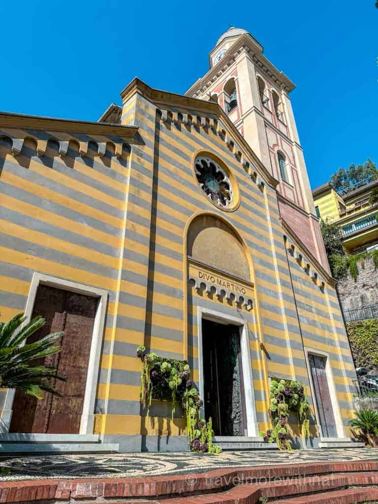 Striped yellow and gray facade of Chiesa di San Martino church with decorative floral displays and a bell tower. This is a must-see stop during a day in Portofino.