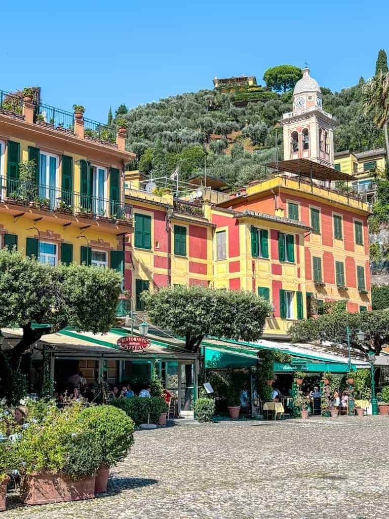 Colourful buildings with green shutters and awnings line a paved plaza with outdoor dining areas in a sunny Italian town. This is Portofino's main square and marina area.
