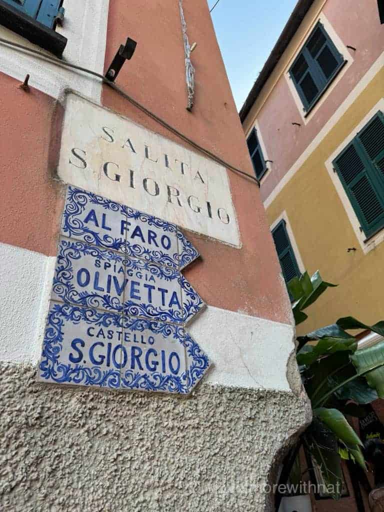 Italian street signs with directions to Al Faro, Oliveta, and Castello S. Giorgio, on a peach-coloured building.