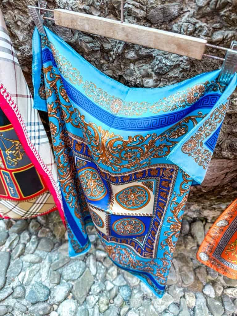 A light blue silk scarf with gold and blue baroque-style patterns, hanging on a wooden hanger outdoors on a stone surface. The perfect souvenir from a day in Portofino!
