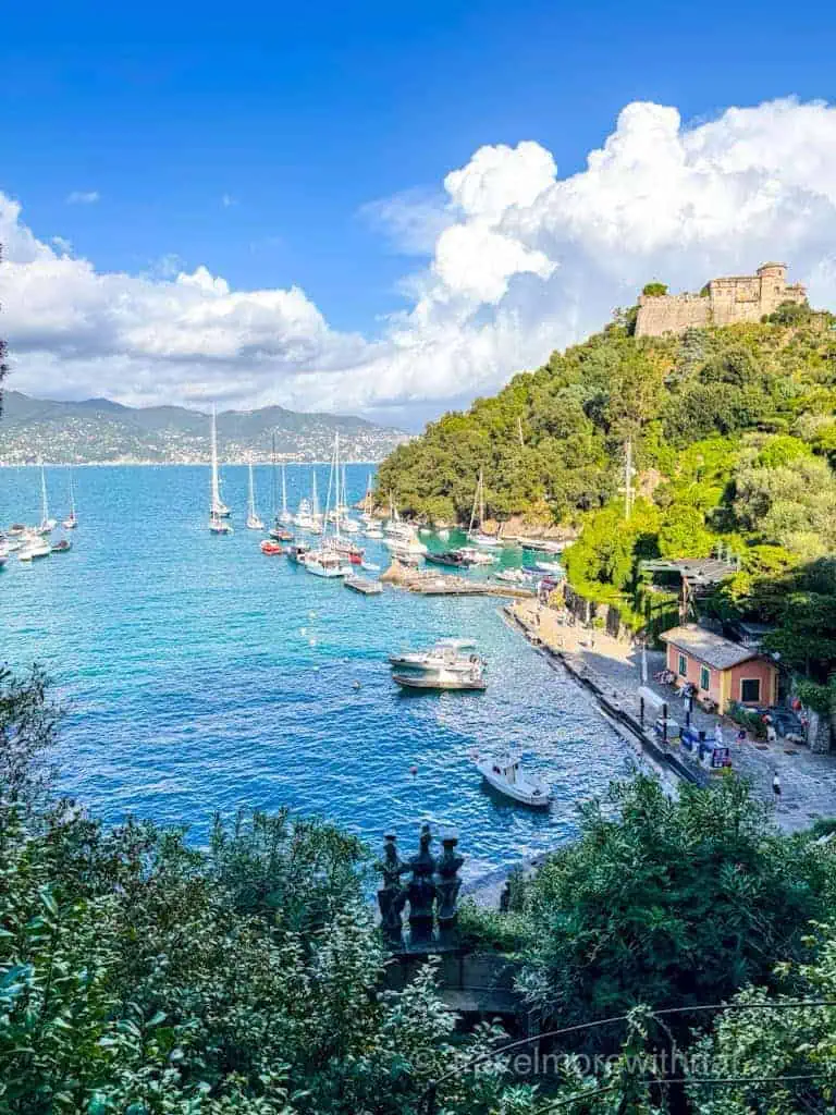 A panoramic view of Portofino harbour with numerous sailboats, a hillside fortress, and lush greenery.
