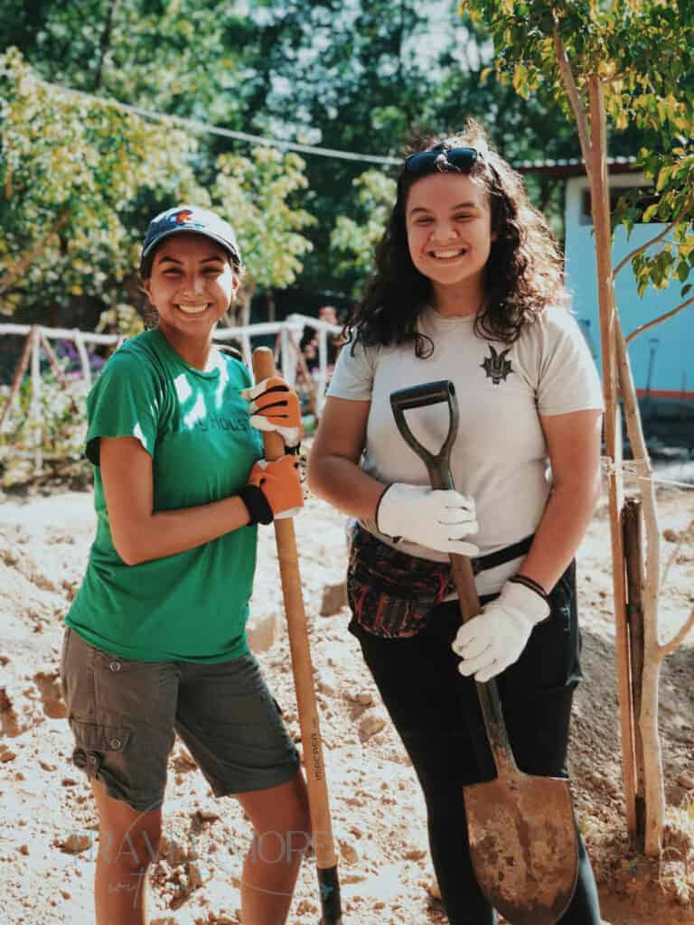 Two women wearing gloves and holding shovels smile while volunteering on a sunny day, surrounded by trees and garden beds. This image illustrates the rewarding experience of volunteering accommodations, offering community and purpose for solo female travellers.