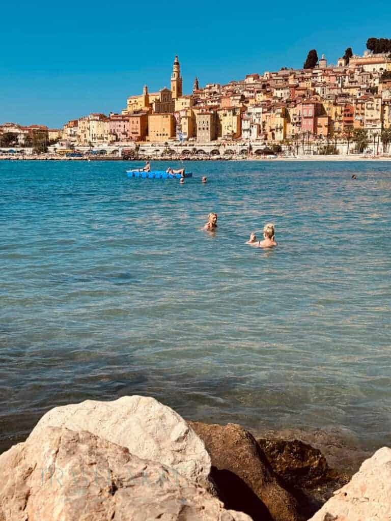 Swimmers in the sea with Menton's colourful hillside buildings in the background - a must-do when visiting this French coastal town.