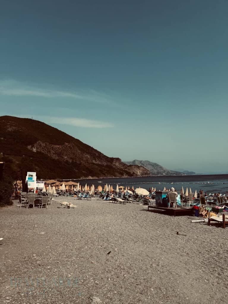 Pebble beach in Budva lined with sun loungers and umbrellas with mountains in the background — A relaxing choice on the list of what to see in Budva for beach lovers.