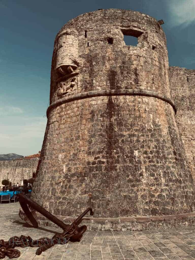 Ancient stone fortress wall in Budva with large iron anchors resting on cobblestone pavement under a bright blue sky — A historic landmark often included in lists of things to do in Budva.