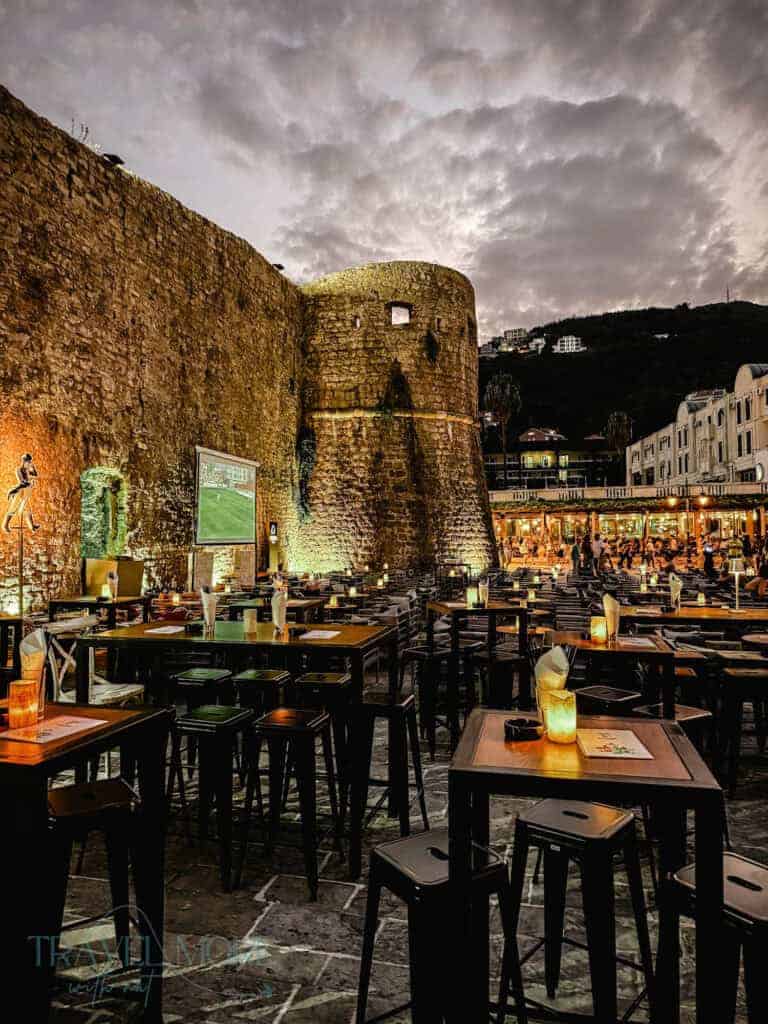 Outdoor dining tables lit by warm lanterns against the illuminated walls of Budva’s old town at dusk — A scenic place for dinner and a top experience on a things to do in Budva itinerary.