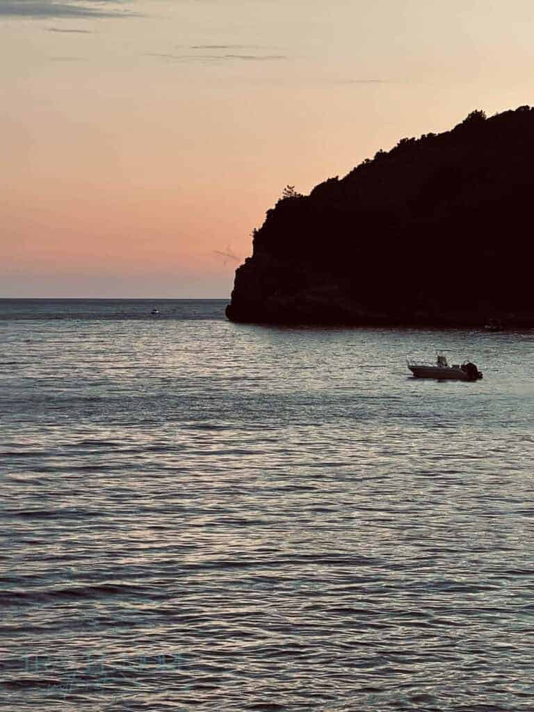 Small boat floating near a rocky coastline at sunset with the sky painted in warm hues — A peaceful scene and a romantic highlight among things to do in Budva.