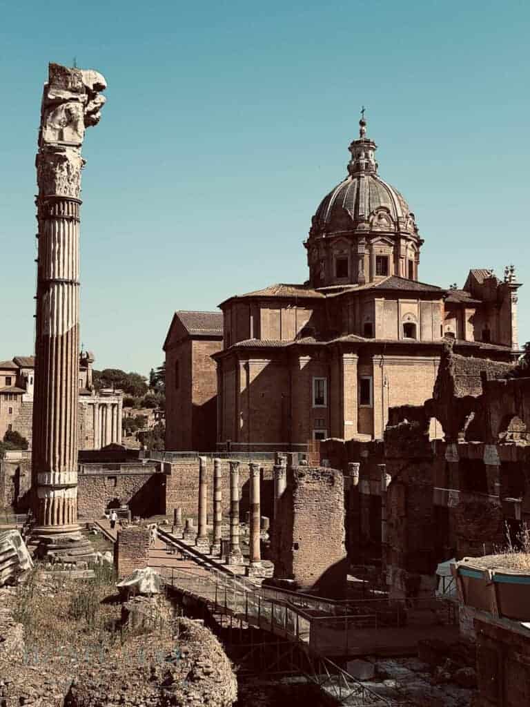 Ancient ruins with tall Corinthian columns and a partially intact brick structure stand in front of the Baroque dome of the Church of Santi Luca e Martina in the Roman Forum, under a clear blue sky.