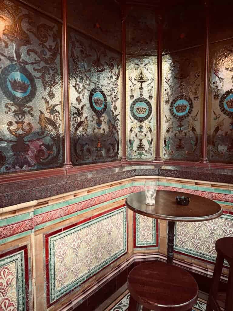 Close-up of a corner booth inside The Crown Liquor Saloon, showcasing its ornate Victorian tiled walls with intricate crown and floral patterns. A small round table holds an empty glass and ashtray.