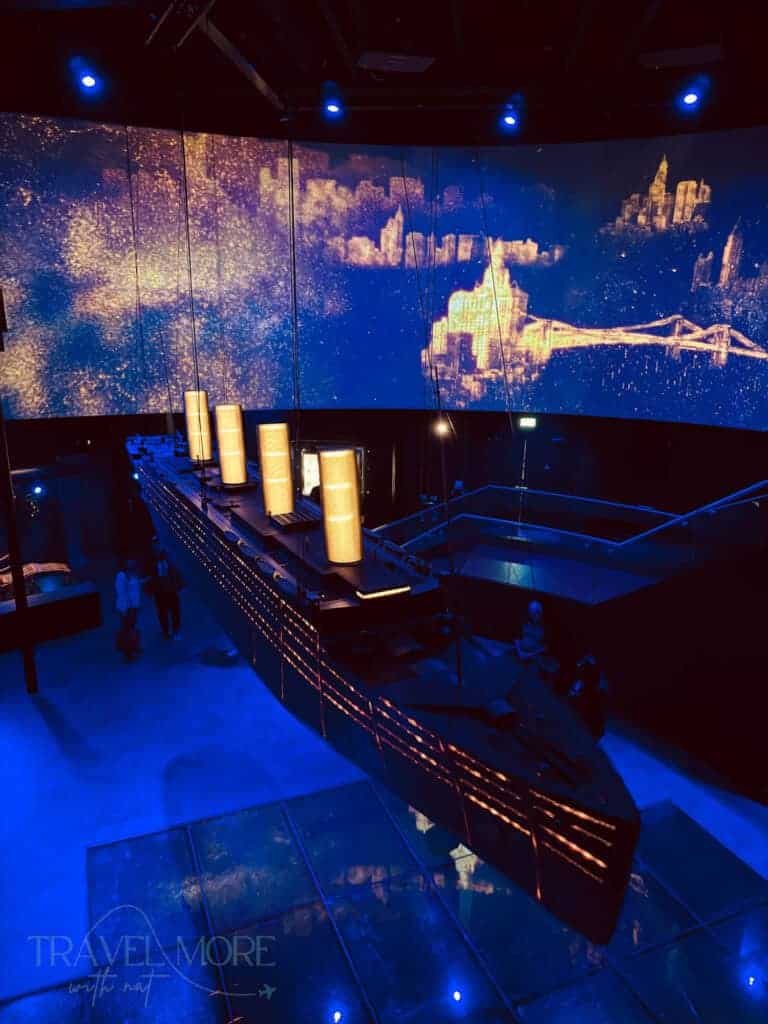 Interior display at Titanic Belfast showing a suspended illuminated model of the Titanic ship above a glass floor. The curved screen in the background displays glowing cityscapes under blue lighting.