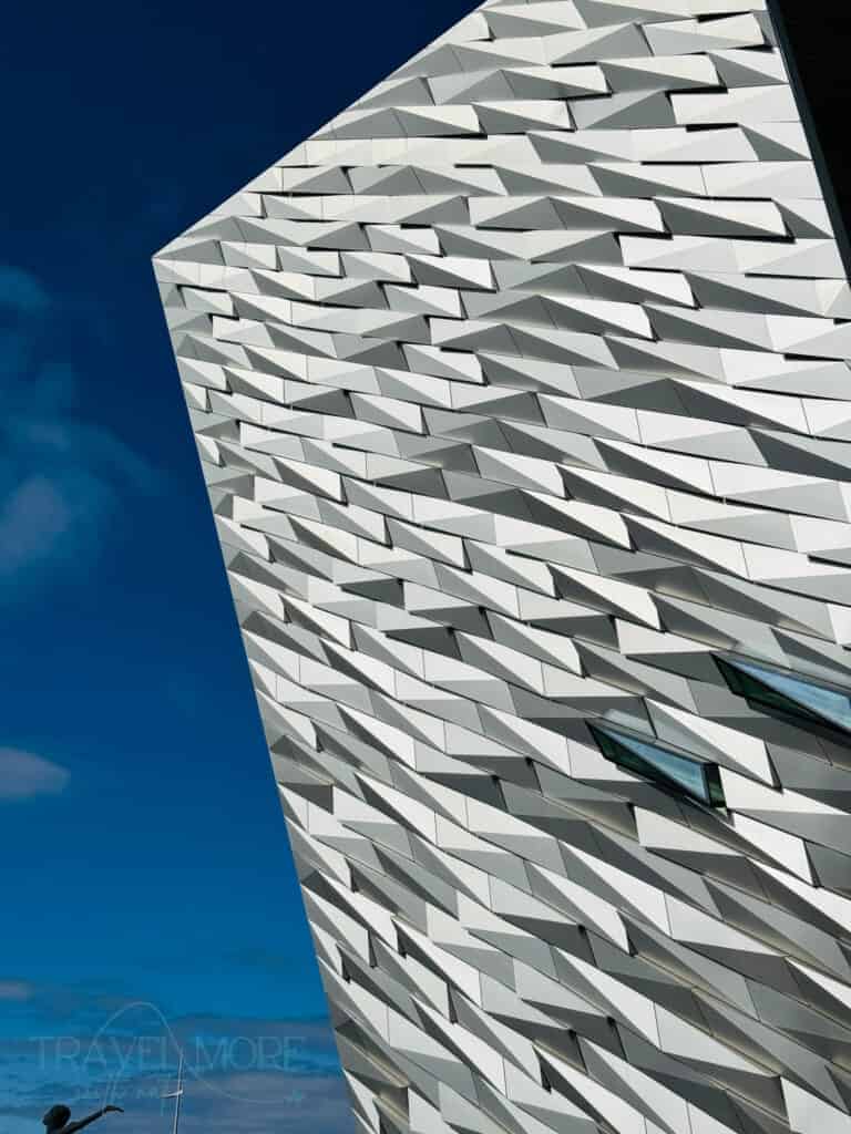 Close-up of the angular facade of Titanic Belfast museum, designed to resemble a ship's hull with metallic, shard-like panels. The dramatic architecture stands against a bright blue sky.