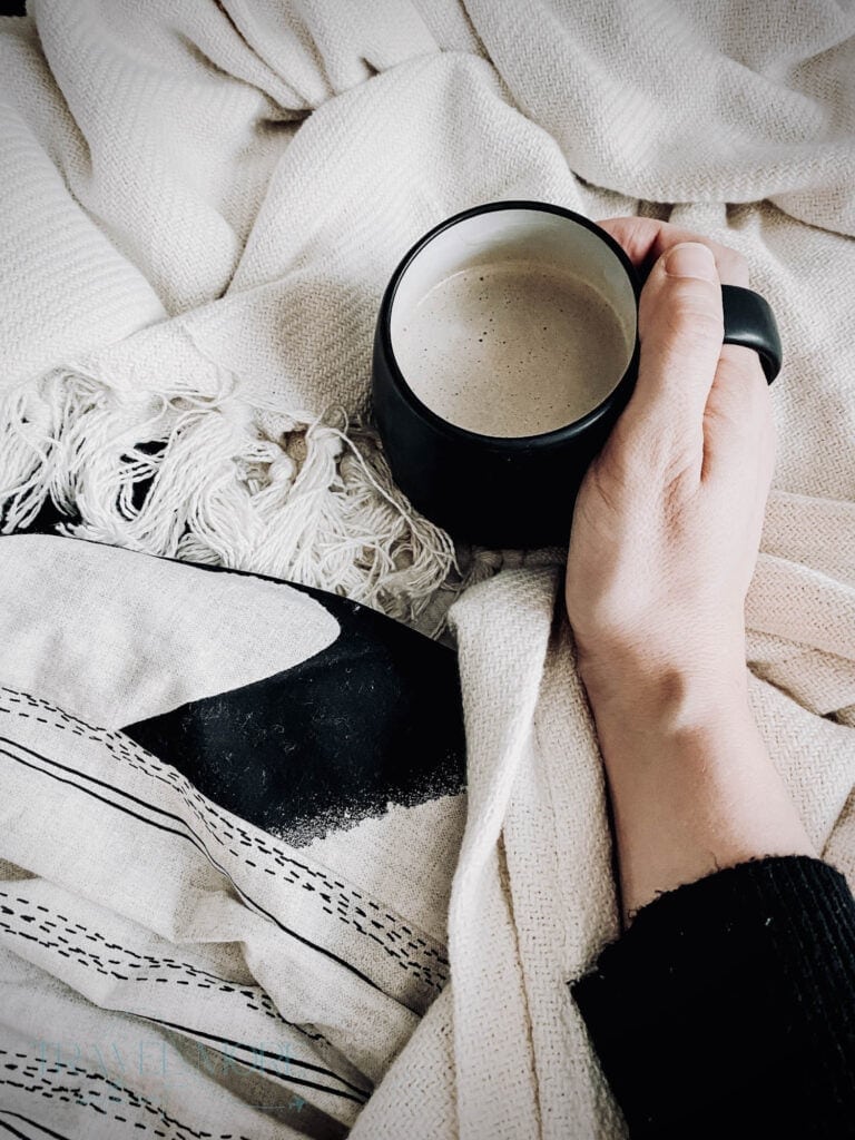 Close-up of a hand holding a chamomile tea mug while wrapped in soft blankets, evoking a cozy, early bedtime routine. A relatable moment for early flight tips that emphasize comfort and restful before heading to the airport.