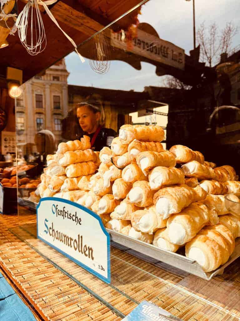 Pastry stall stacked with cream filled pastries labeled "Ofenfrische Schaumrollen" while a vendor works behind the counter. Sweet Austrian treats like these are popular snacks to try at Vienna Easter markets.
