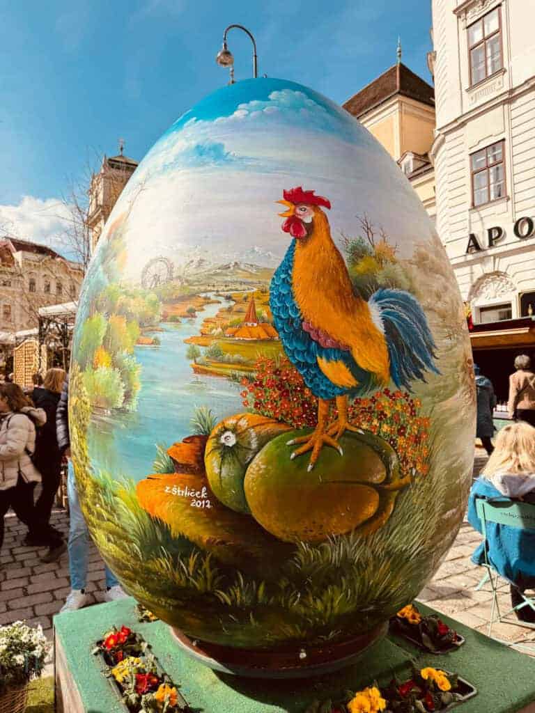 Giant painted Easter egg featuring a colorful rooster and countryside landscape displayed in a busy city square with visitors walking past. Large themed displays like this help decorate the Vienna Easter markets each spring.