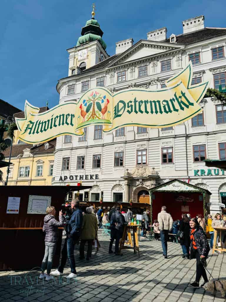 Crowds browsing wooden market stalls beneath a banner reading "Altwiener Ostermarkt" in front of a historic building in Vienna. This traditional square hosts one of the most famous Vienna Easter markets.