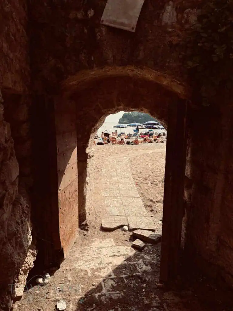 Rocky passage opening onto a sunny Budva beach, with a tiled path leading to the sand, dotted with sun loungers and people.