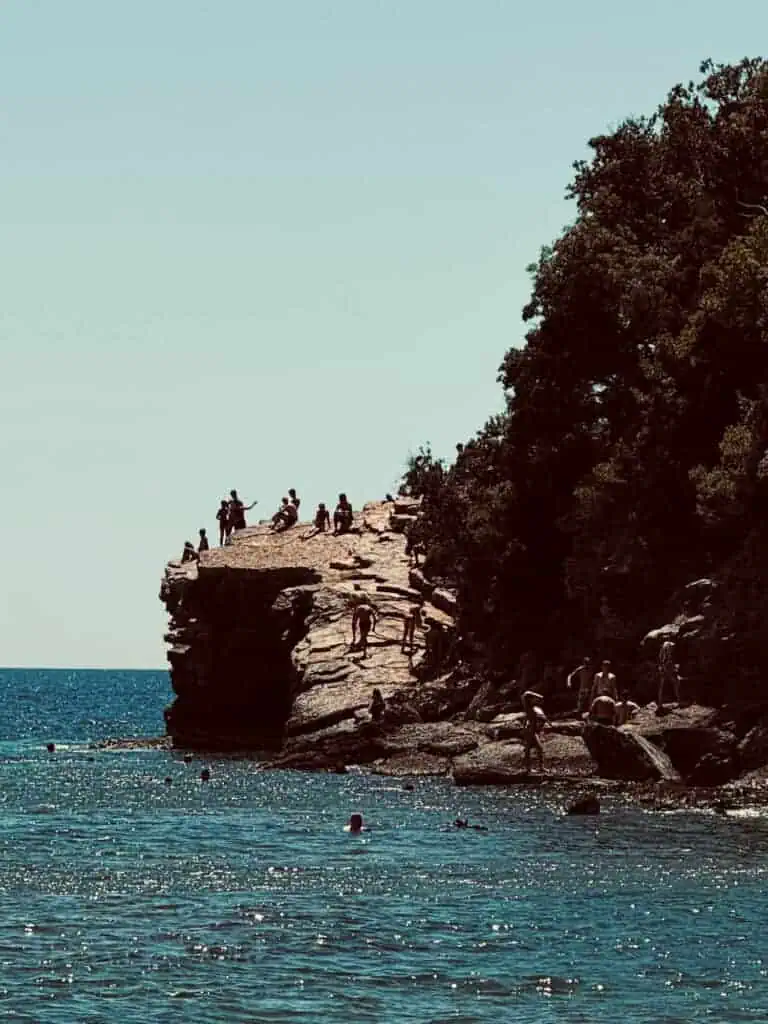 Swimmers and sunbathers along the rocky beaches of Budva, with clear blue water and a steep cliff.