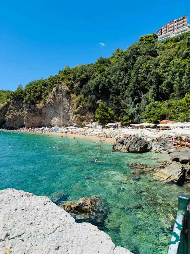 Clear turquoise water at one of the Budva beaches, with a packed pebble cove and steep green cliffs.