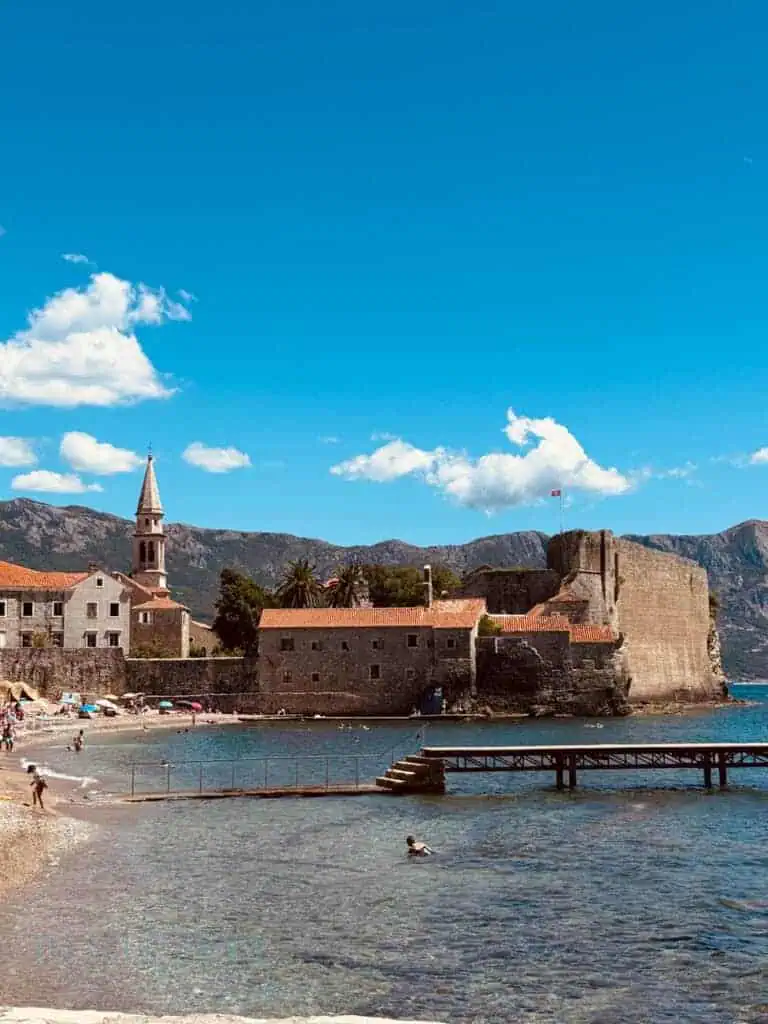 Budva beach with a stone fortress, church tower and calm clear water.